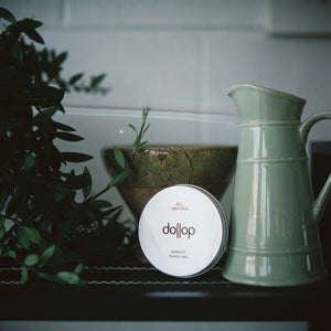 Shelf with tin of dollop pomade surrounded by lush plants and green pottery bowl and jug